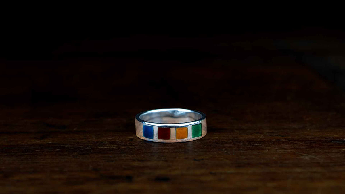 A silver band ring featuring a row of small rectangular inlay enamels in blue, red, yellow, and green, resting on a dark wooden surface.